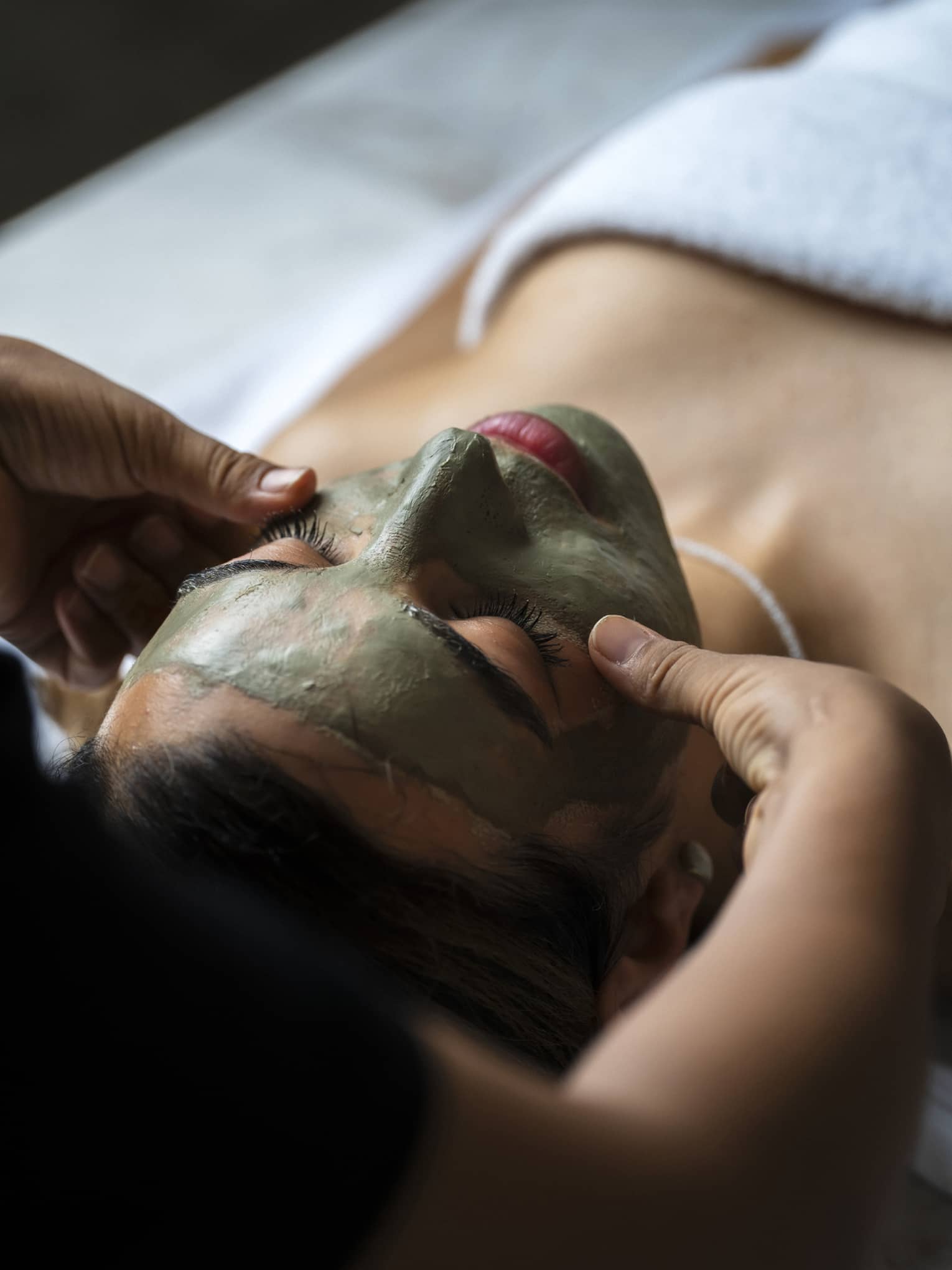 A person lying supine on a spa treatment table receives a facial treatment.