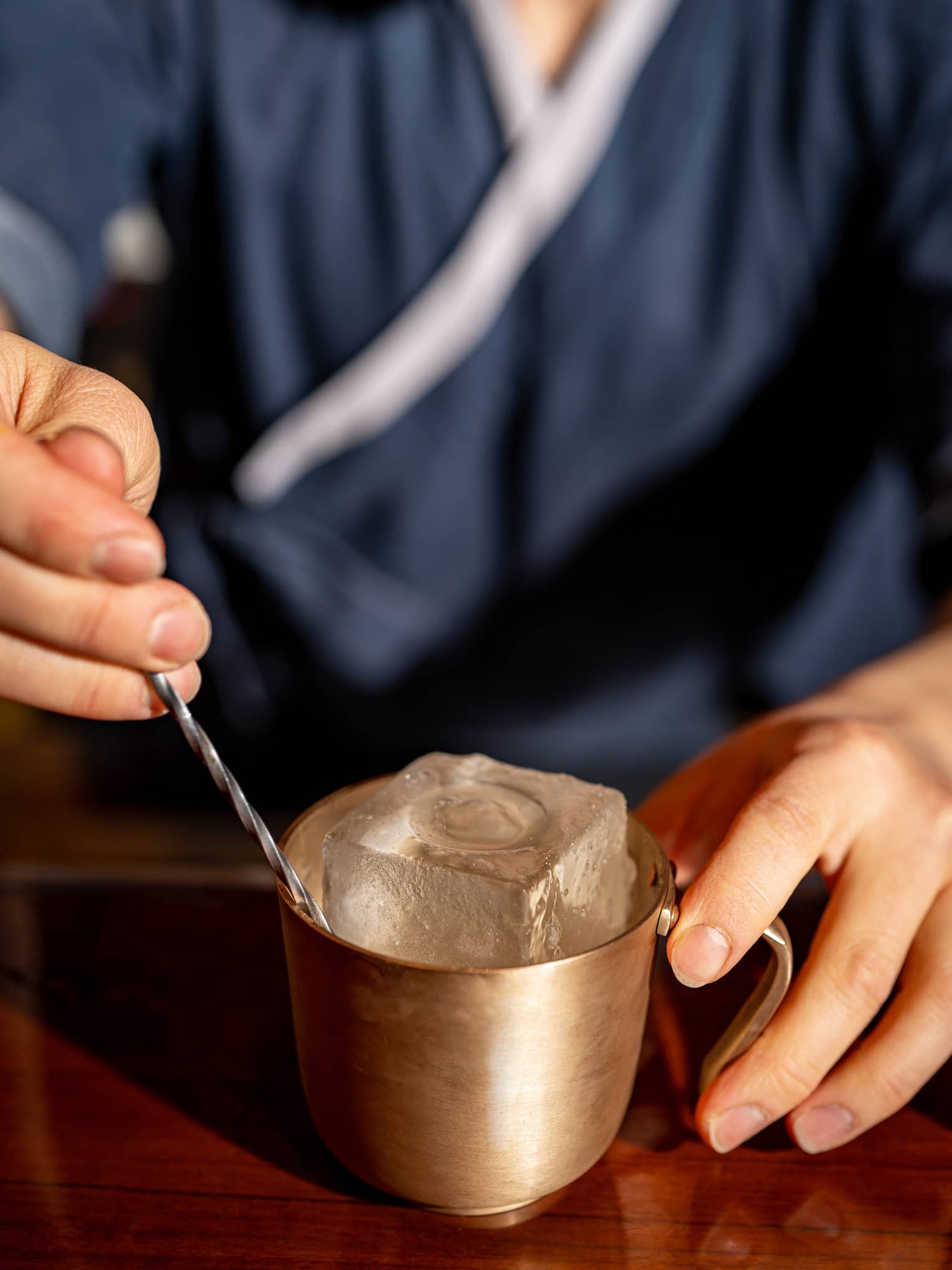 Close-up of a hand on a gold metallic mug containing a giant embossed ice cube and a twisting metal stir stick.
