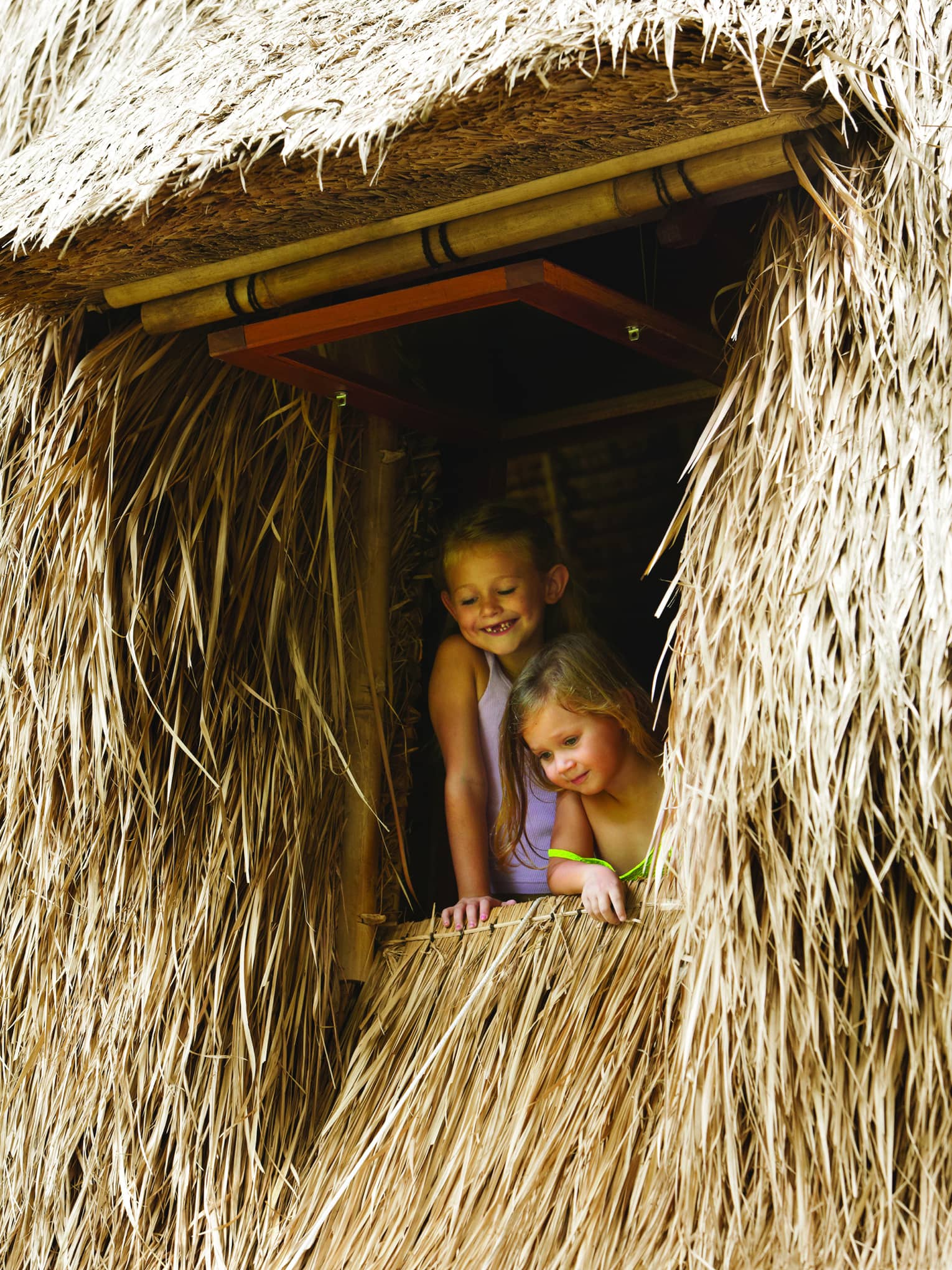 Two smiling young children look out from window of thatched building