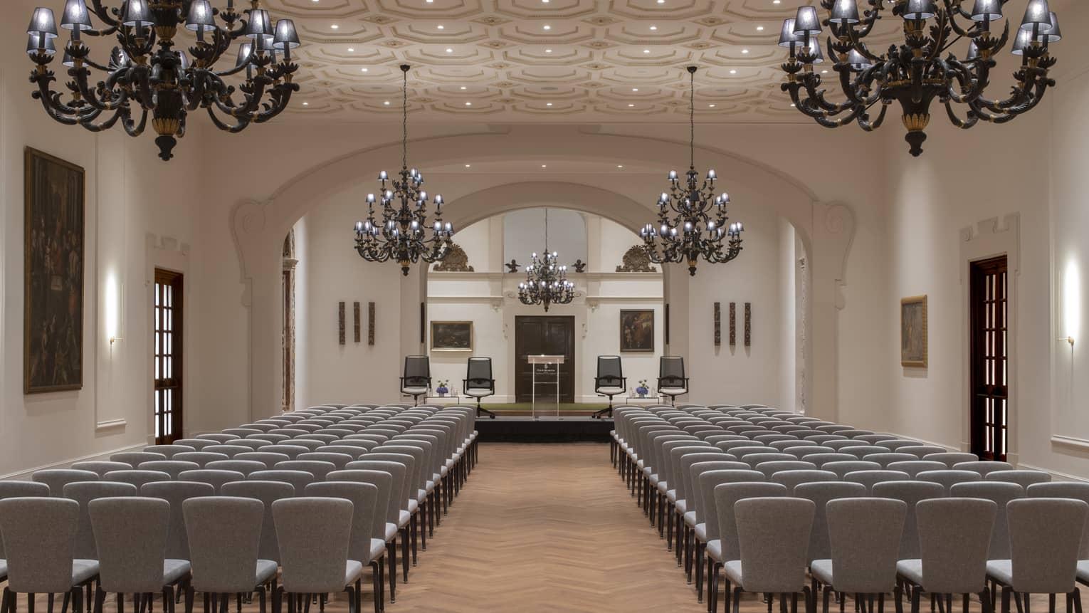 A spacious, neutral-toned space with rows of chairs, a stage area at one end and a textured ceiling, softly lit by elegant chandeliers, recessed ceiling lighting and wall sconces.