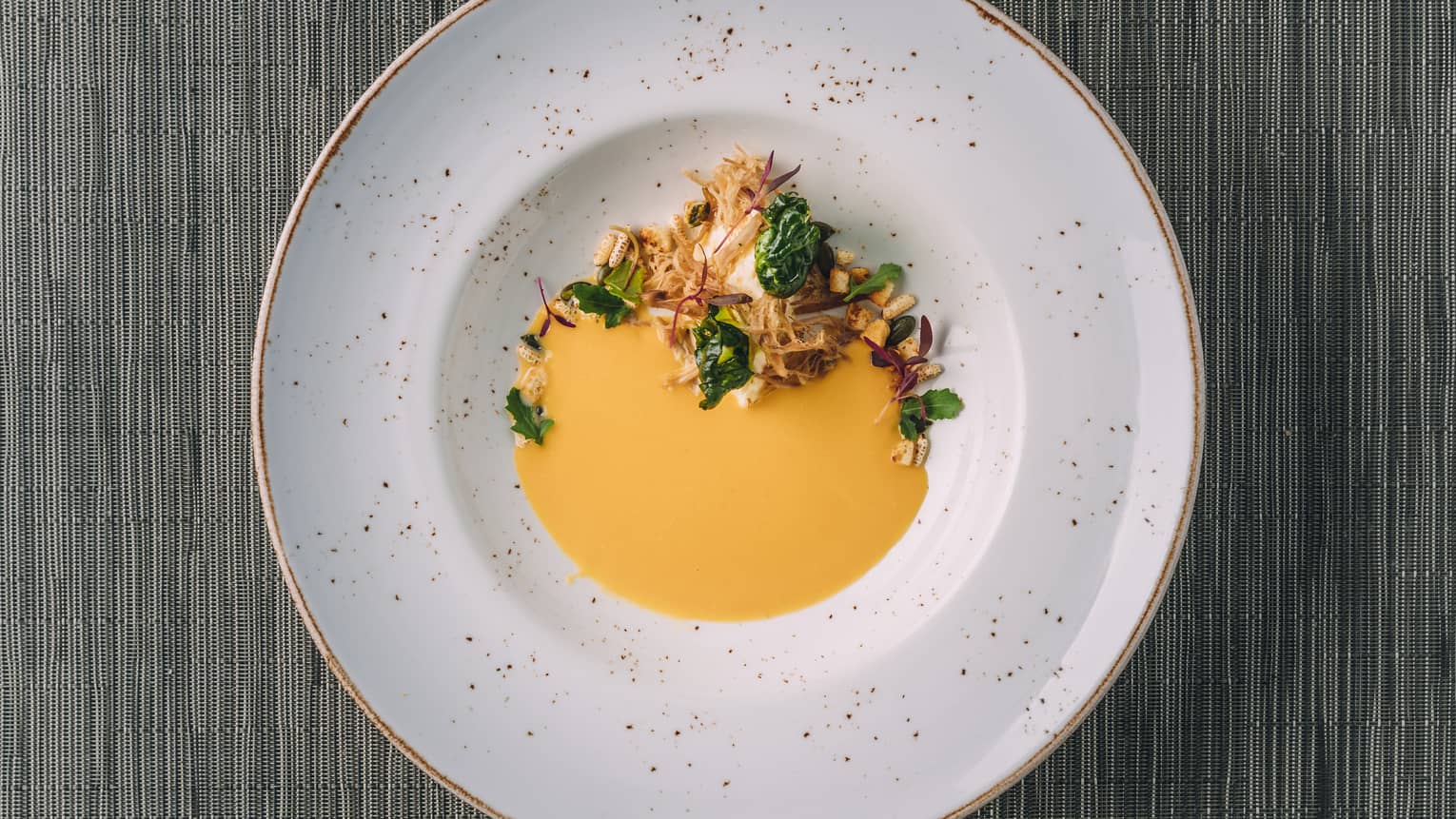 Chilled yellow soup with herb garnished in a shallow speckled bowl with gold trim