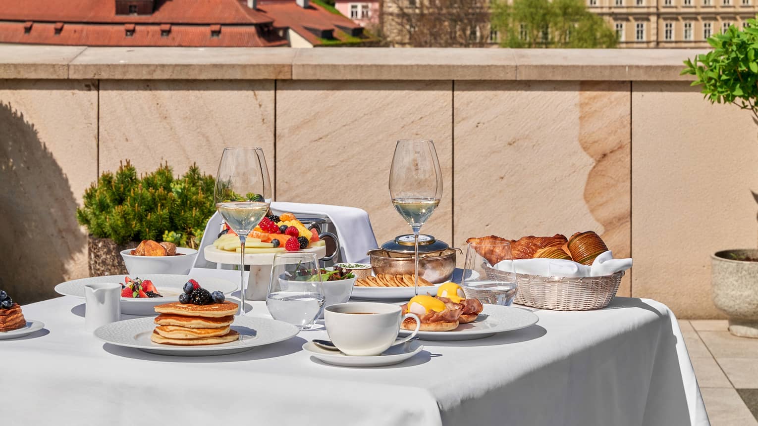 A white cloth?covered dining table on a terrace, set for a meal, with city views in the background