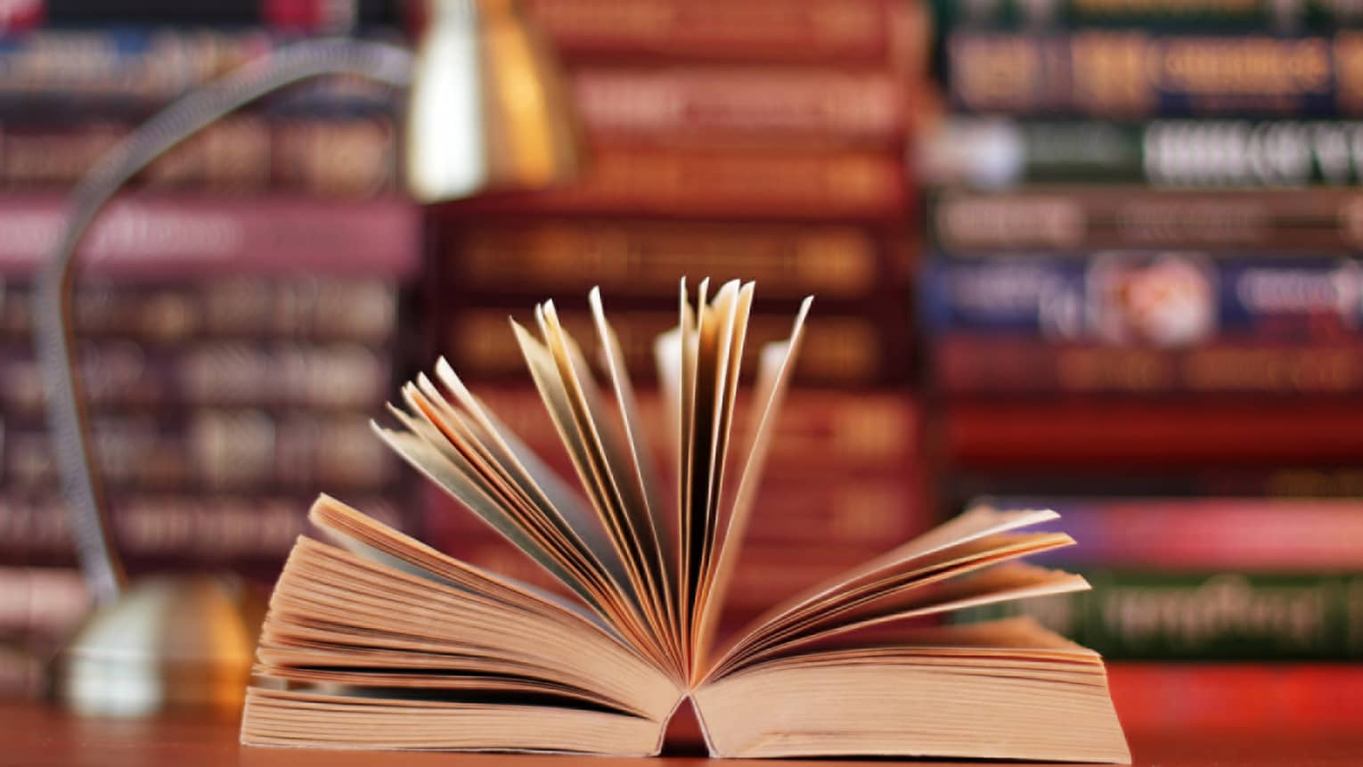A book is opened, with pages fanned, lamp and stacks of books in background 
