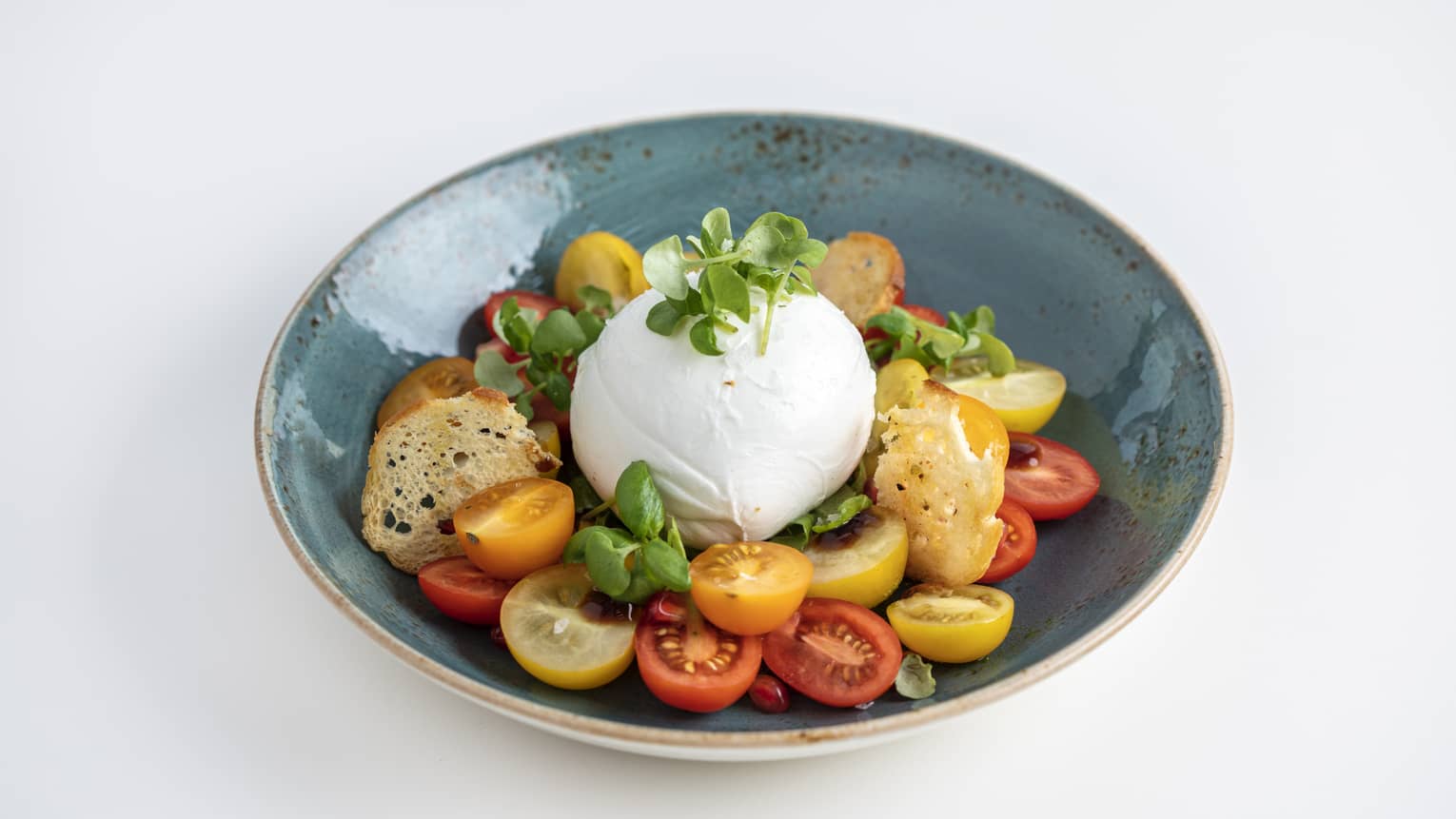 Burrata served with cherry tomatoes, arugula, pomegranate, balsamic drizzle and basil pesto in a round blue bowl