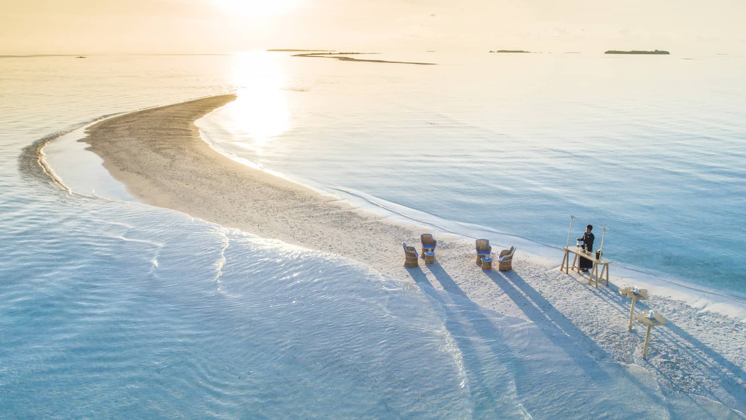 Person stands at rustic wooden bar on strip of sand with chairs, surrounded by water