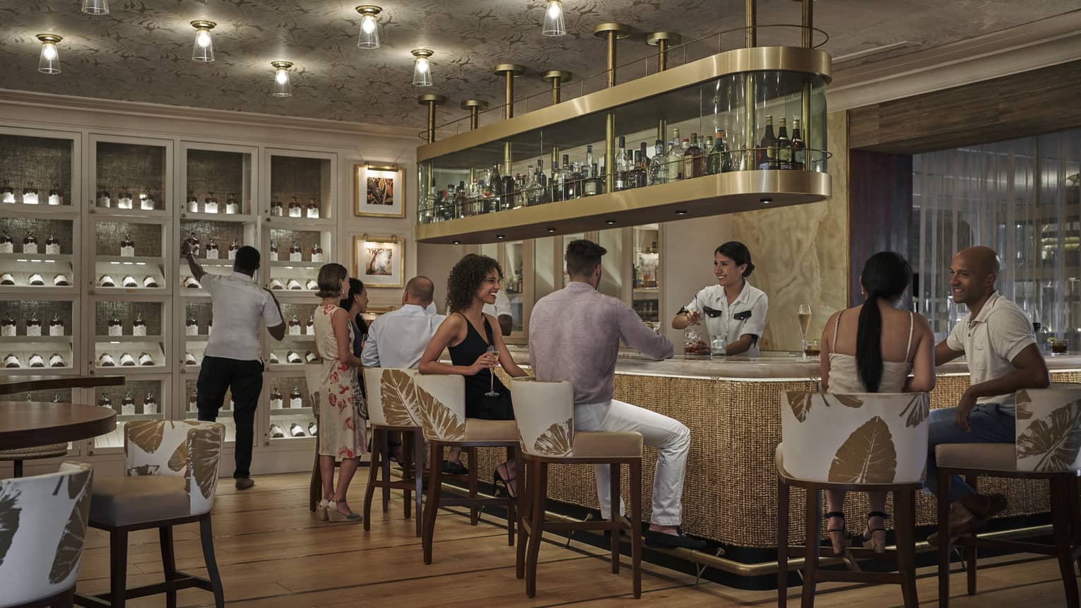 People mingle at a bar, with a hanging shelf holding bottles and a wall of wine shelving