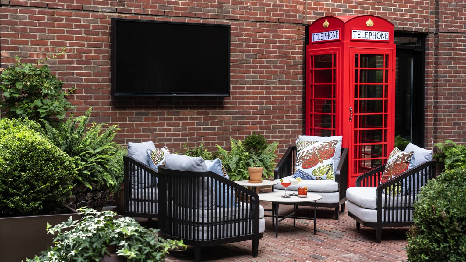 A small sitting area outside surrounded by brick walls, plants, a tv and red phone booth.