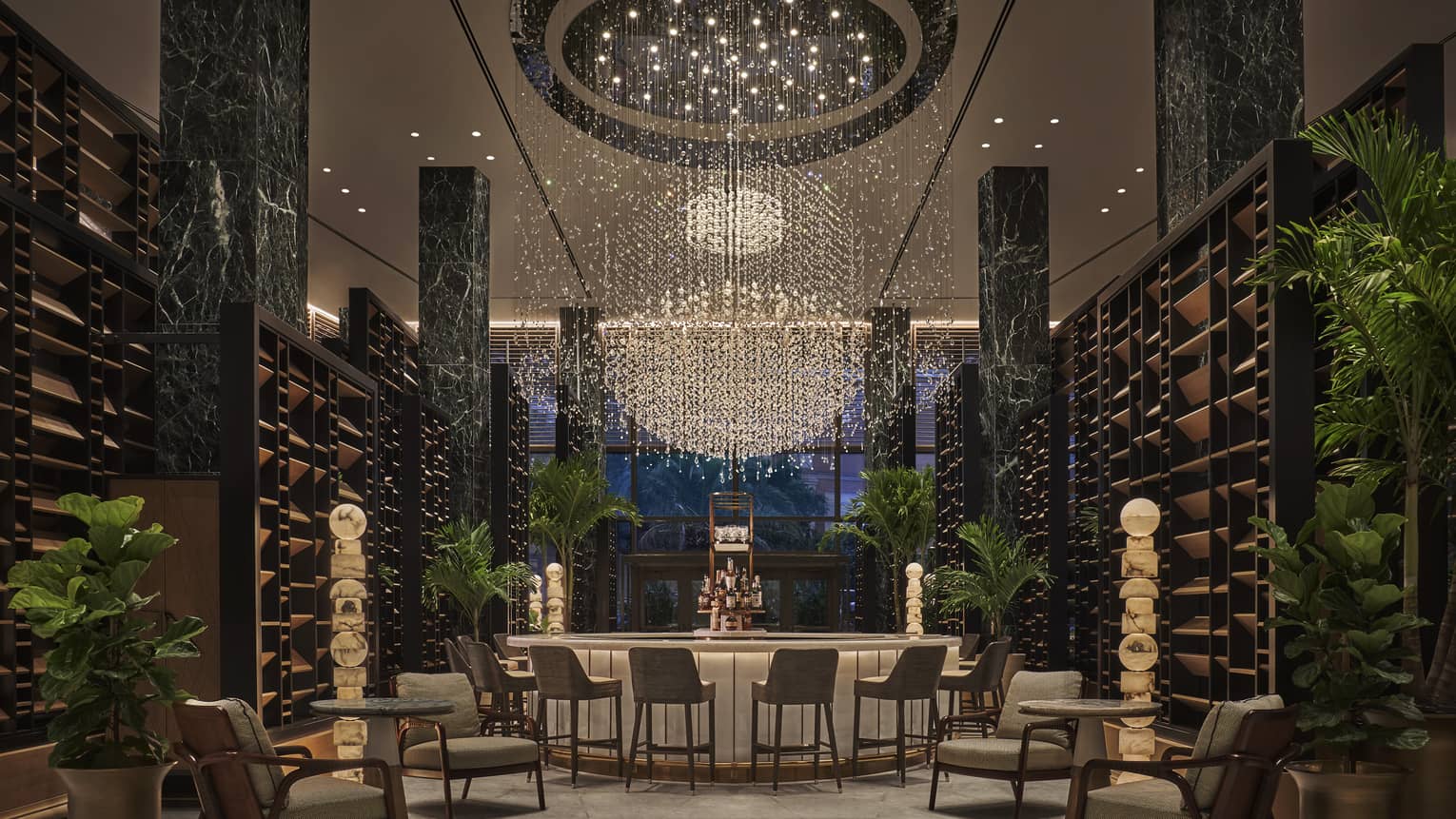 Chandelier Bar with six jade pillars flanked by wooden shelves and potted palms, glass-topped bar in the middle with six cocktail chairs, giant chandelier hanging overhead