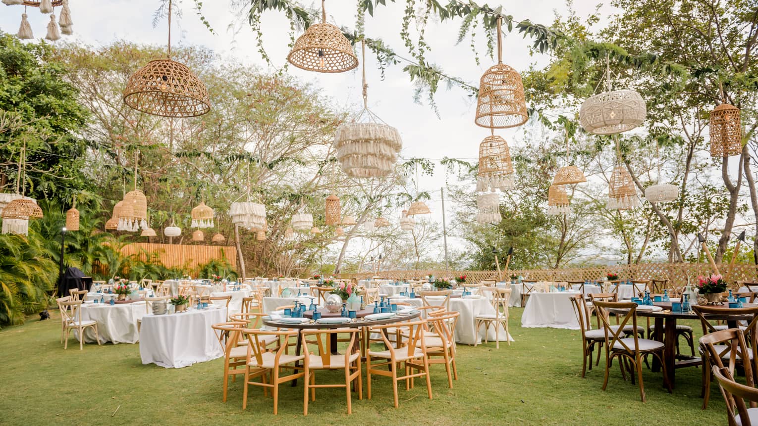 A lawn is set for an outdoor party with various round tables, white table cloths and a variety of straw light fixtures hanging from vined ropes overhead