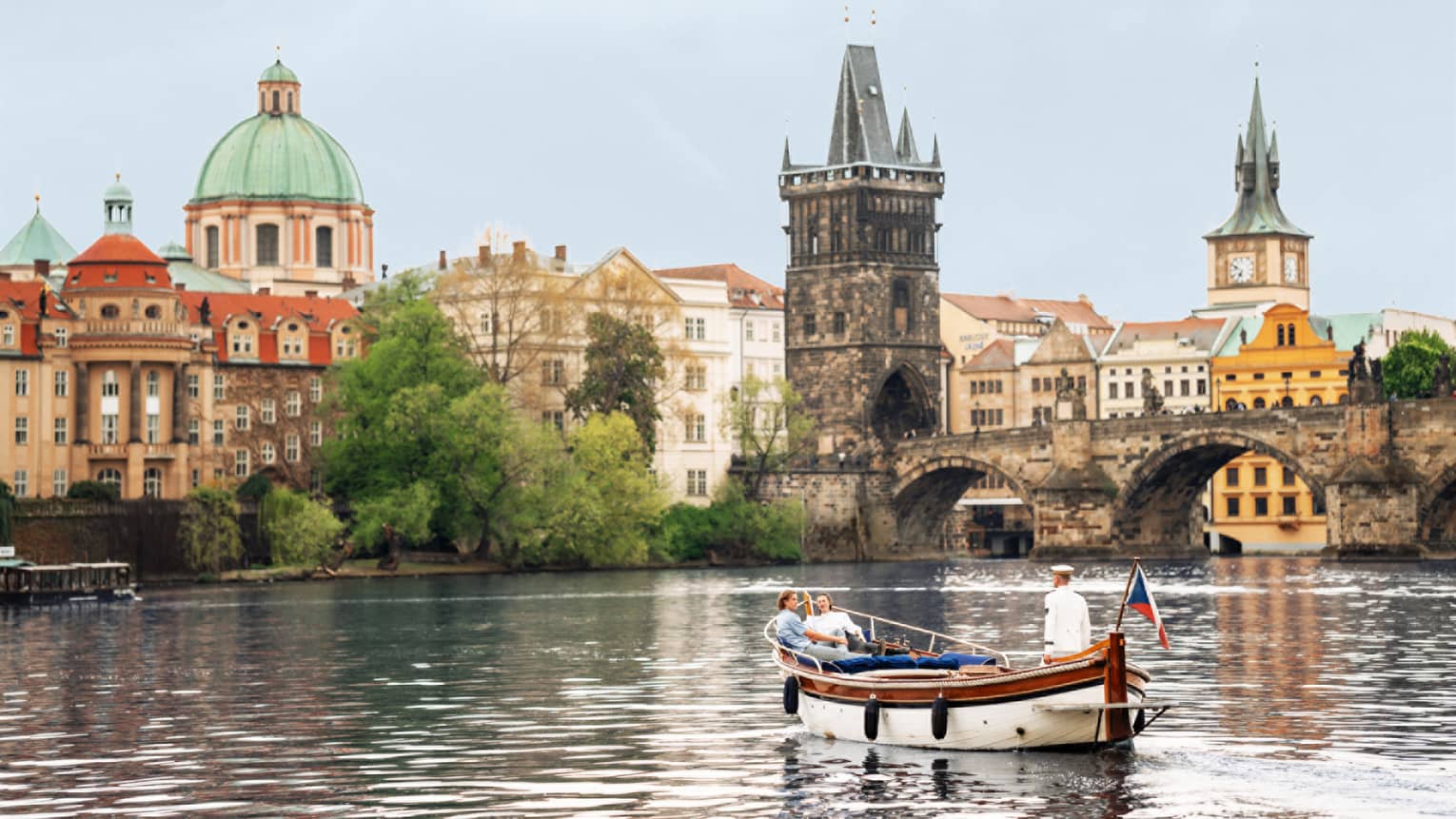 Couple in wooden boat with captain on Vltava River with baroque buildings in backdrop