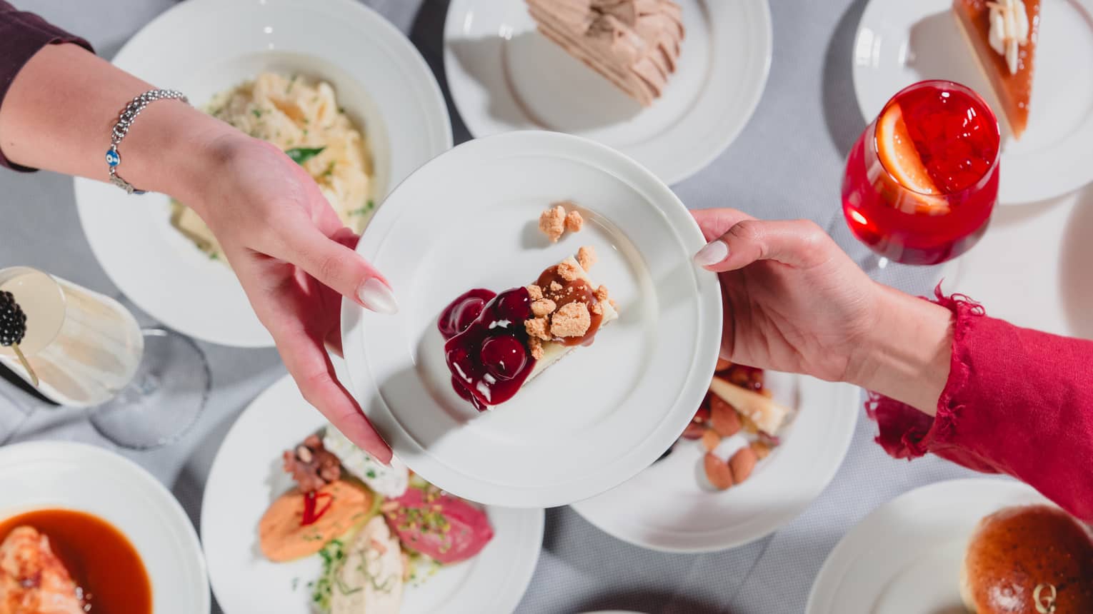 One hand passes along a white plate to another hand with a variety of dishes of food seen below on a table