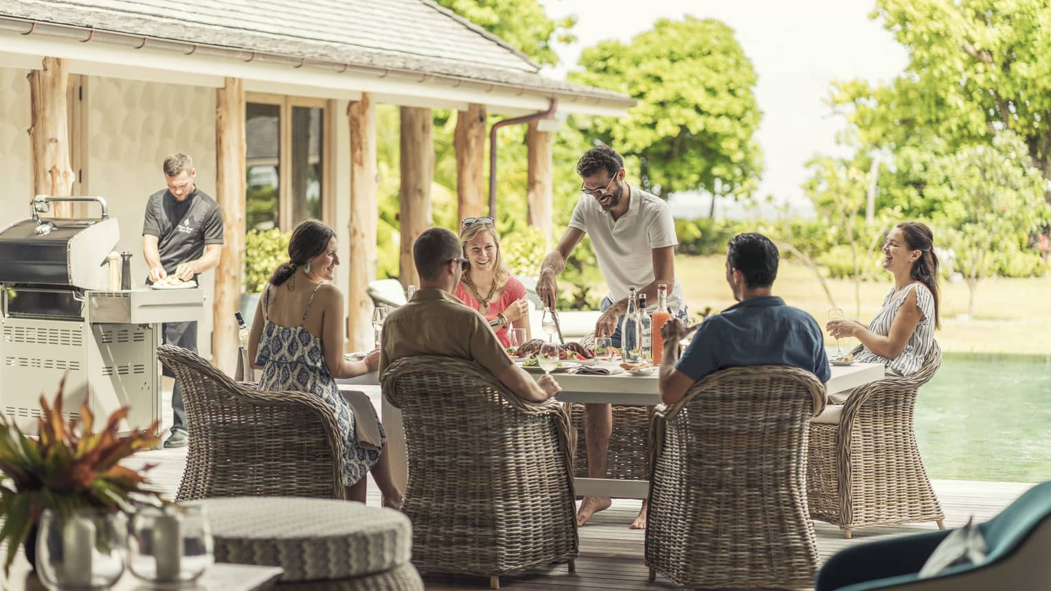 Group of friends enjoying an outdoor meal together on a sunny patio, with a chef preparing food on a grill nearby, creating a relaxed and social atmosphere.