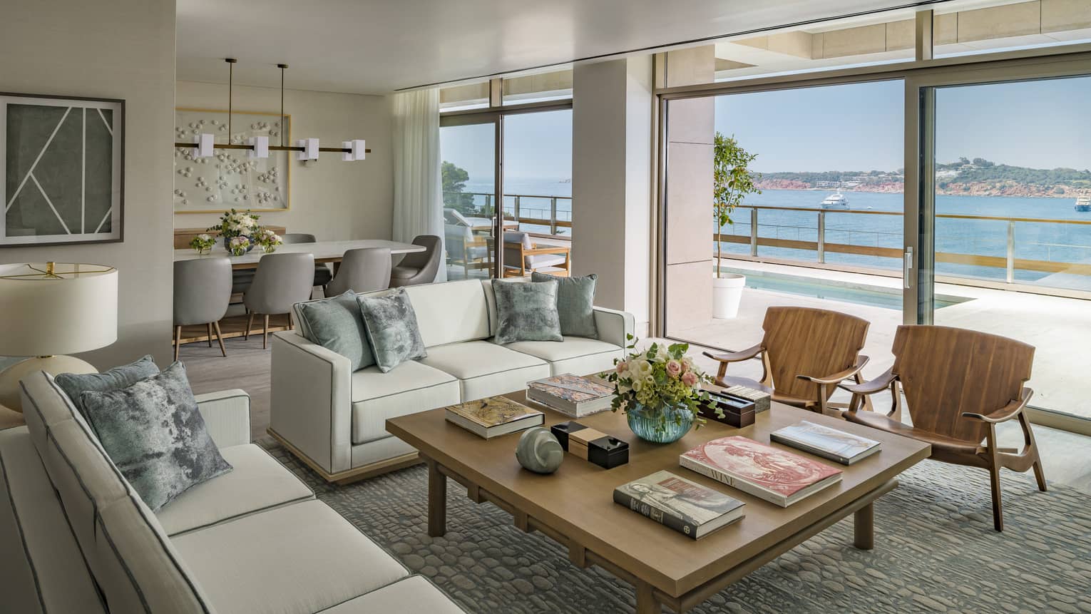 Arion Royal Suite living area with white sofas, square coffee table, arm chairs, dining table in background, floor-to-ceiling window with view of plunge pool and Mediterranean Sea