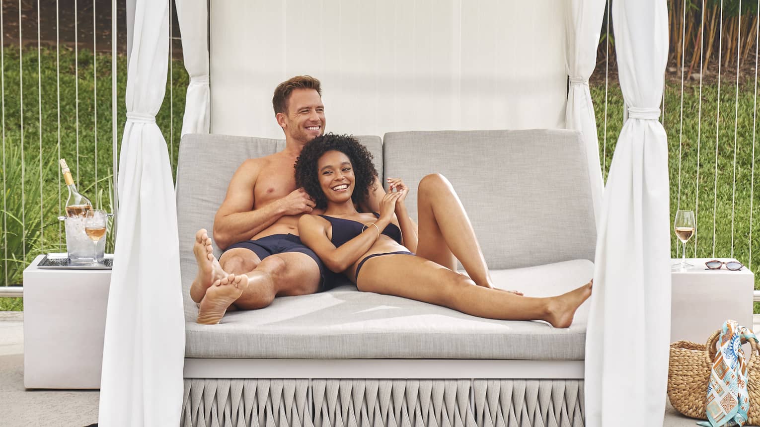 A smiling couple in swimsuits lounges together on a luxurious daybed, a bottle of rosé in a frosty ice bucket beside them.