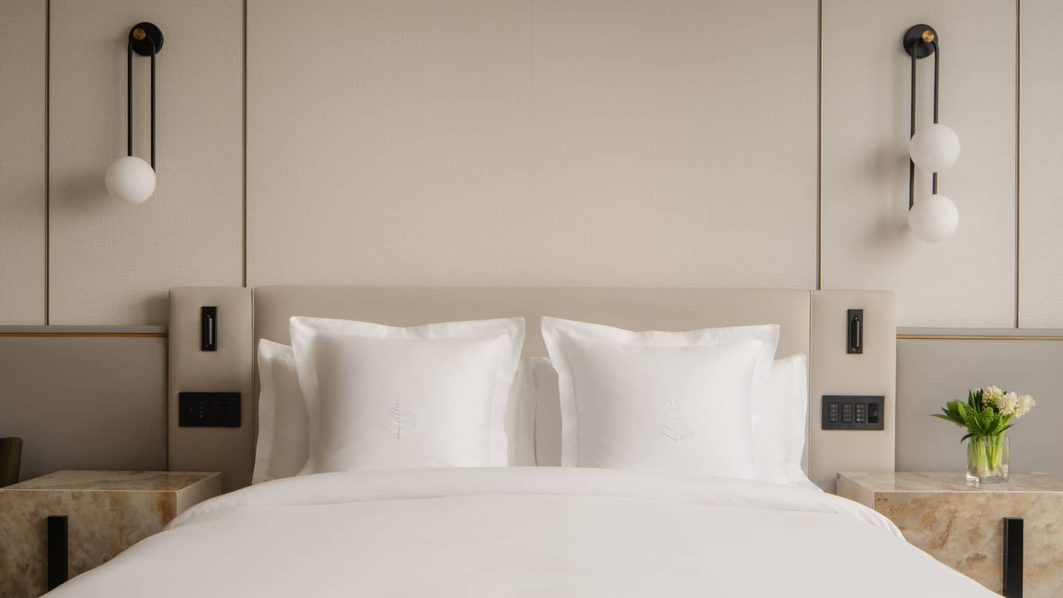 Close-up of a hotel room bed with white pillows, nightstands, bedside controls, a vase with flowers and pendant lights 