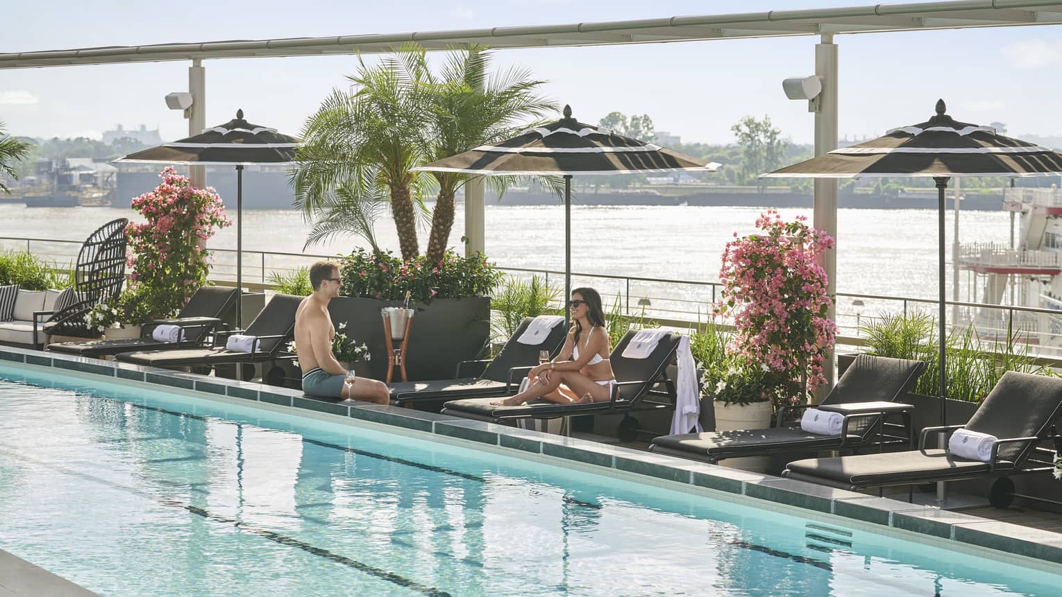 A man and woman lounging next to a pool outside with plants and umbrellas around it.