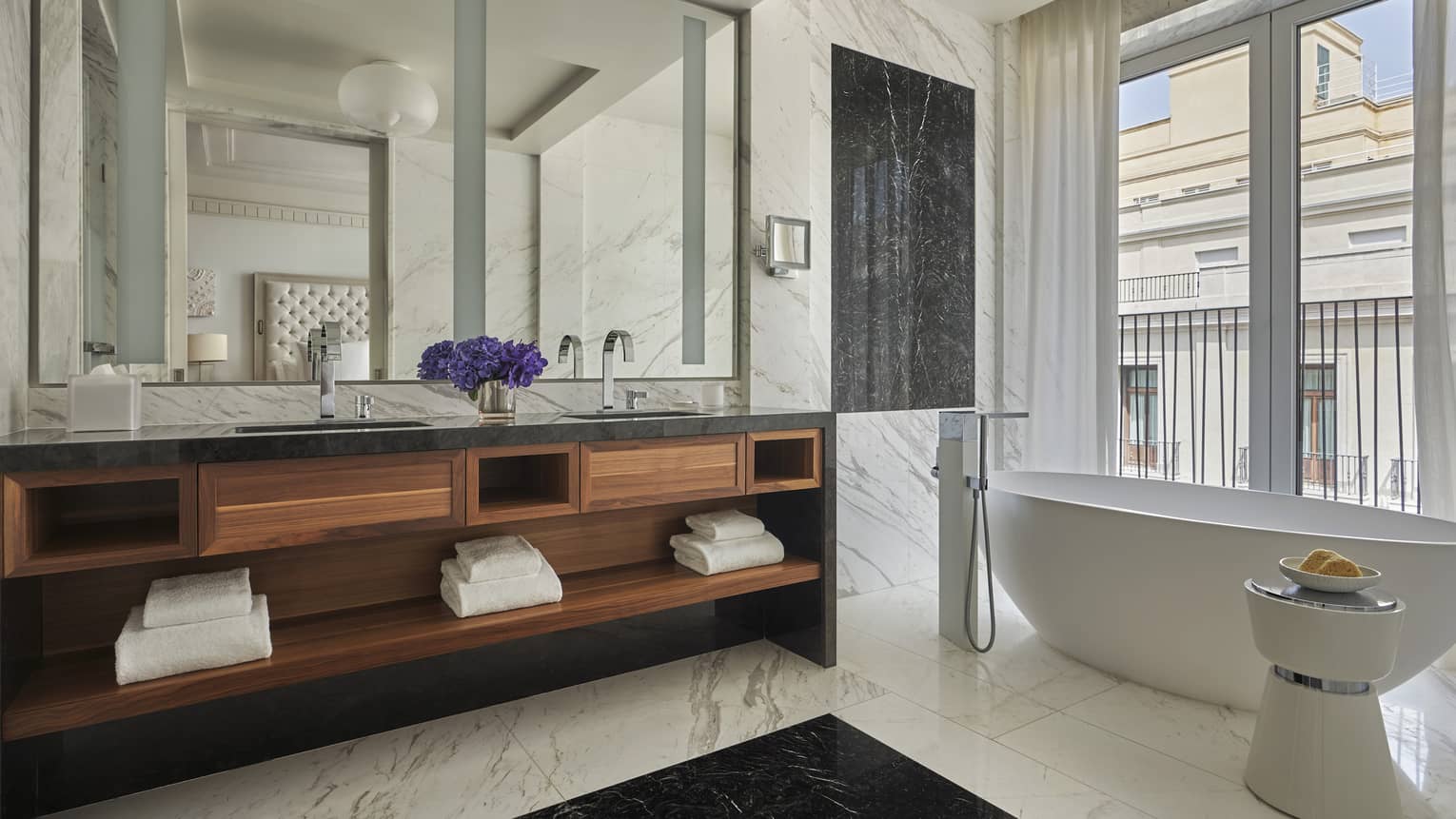 Bathroom with white marble walls and floors, oval white tub, wooden double vanity 