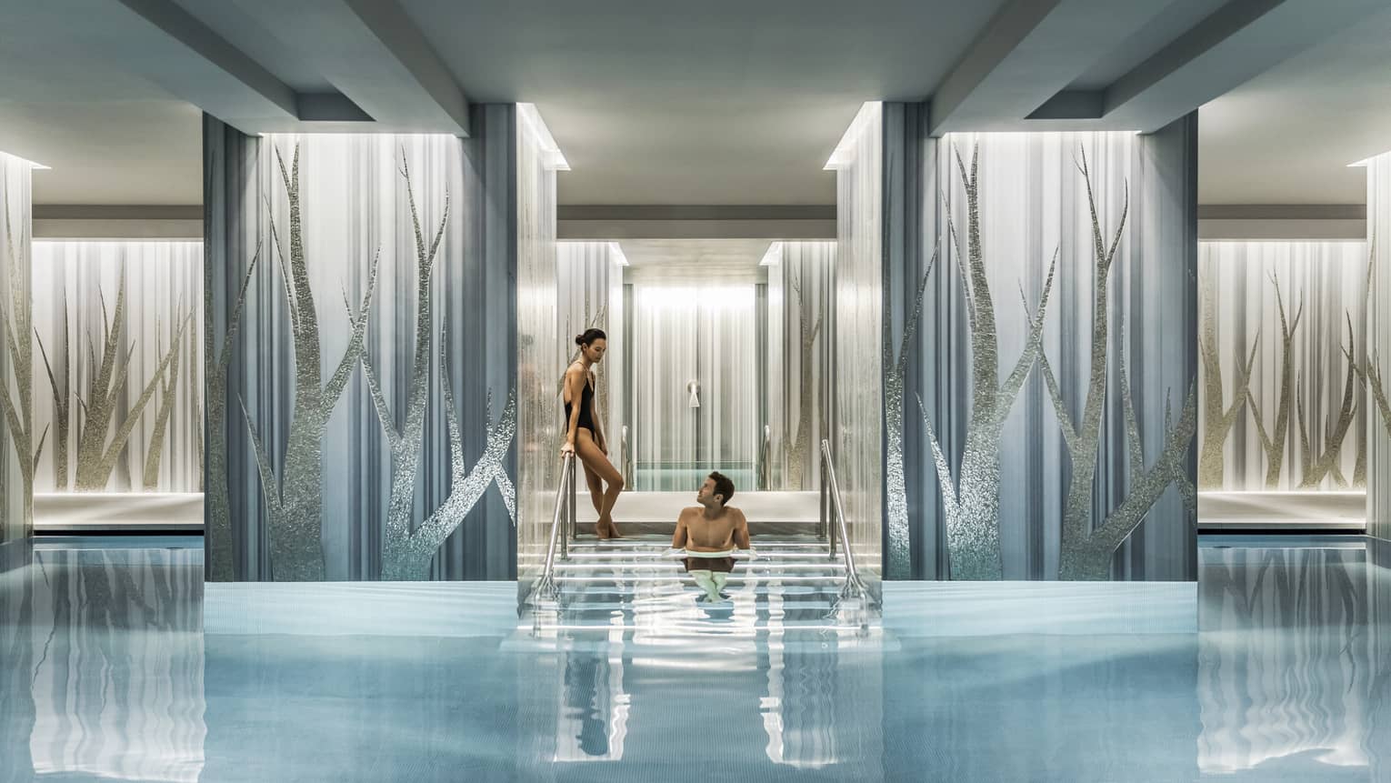 A couple enjoys a serene swimming pool, with one person seated on steps between elegant blue and silver columns.