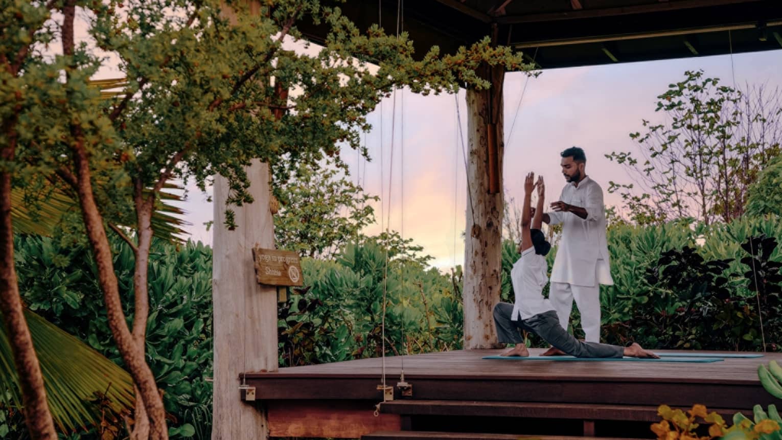 Instructor guides student doing yoga in an open-air pavilion surrounded by greenery