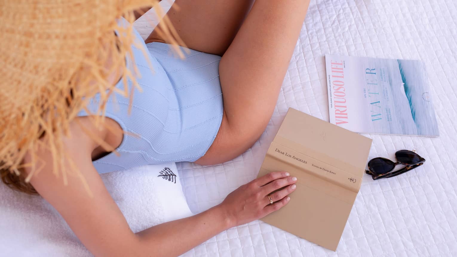 A woman in a blue swimsuit and straw hat holding a tan book while lounging on white bed