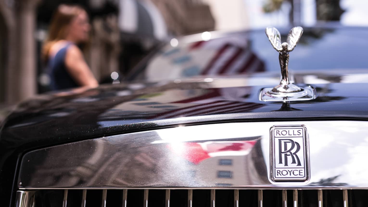 Close-up view of a winged silver ornament on the hood of black Rolls Royce, double-R logo on reflective chrome above grille.