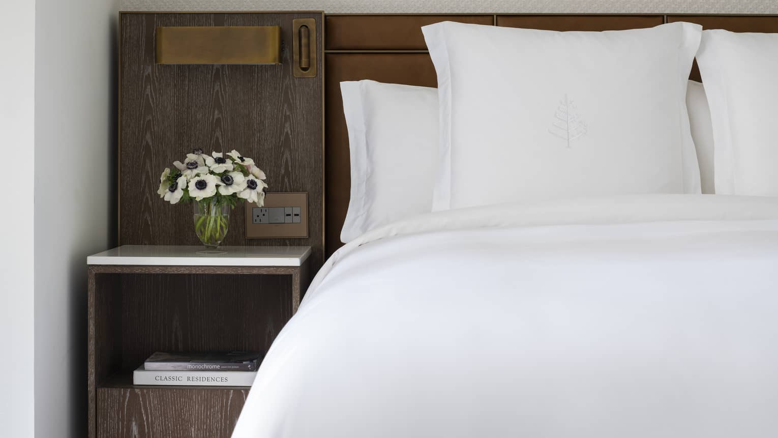 White bed with Four Seasons-embroidered pillows, set next to a wooden night stand with flowers
