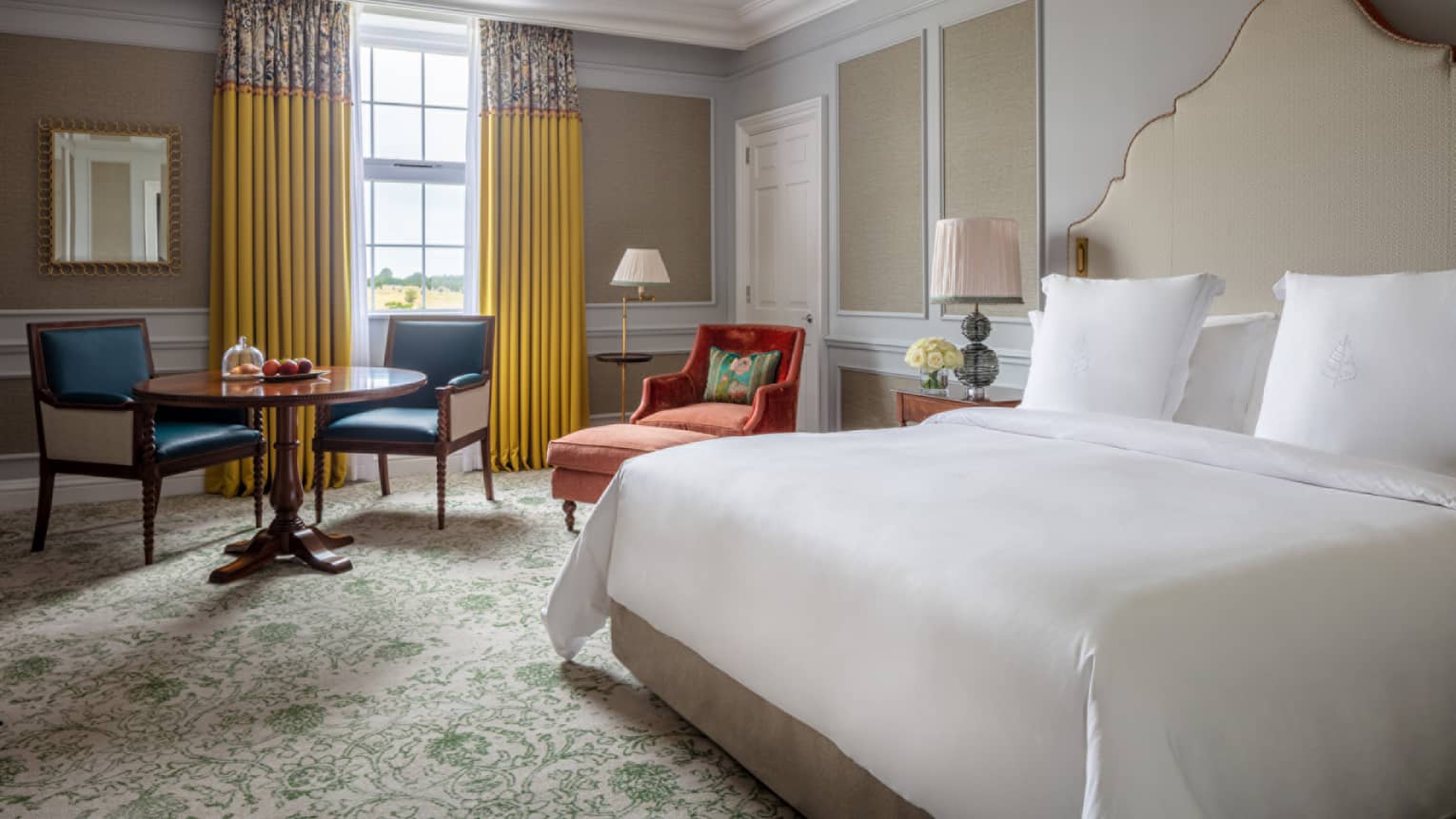 Guest room with king bed and white linens, red chaise, small round table and two chairs, window with yellow drapes