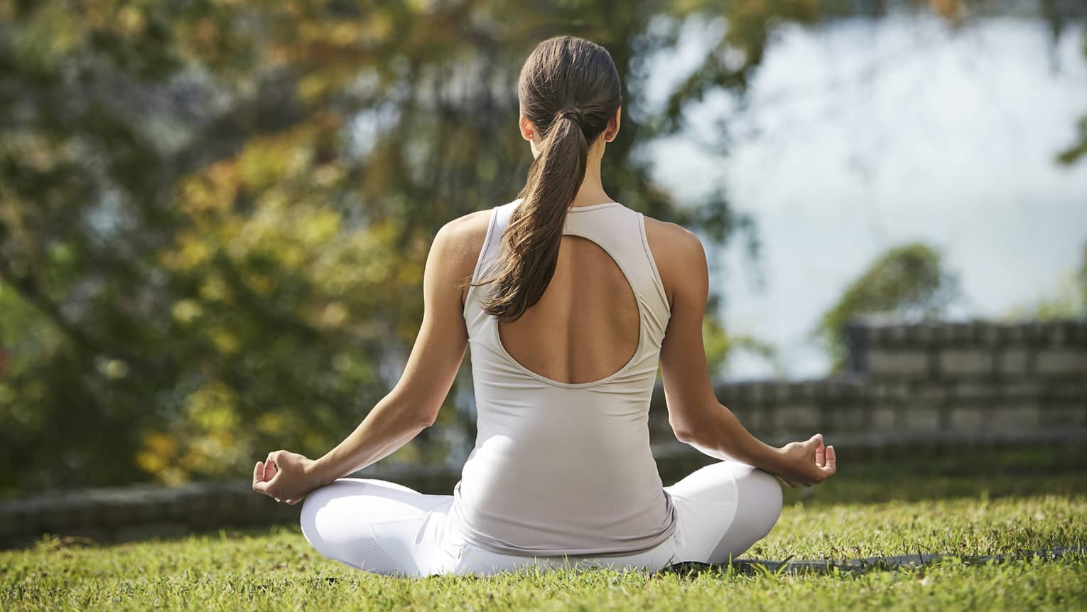 Rear view of a guest in white athletic wear meditating cross-legged with forearms rested on knees on a sunny patch of grass.