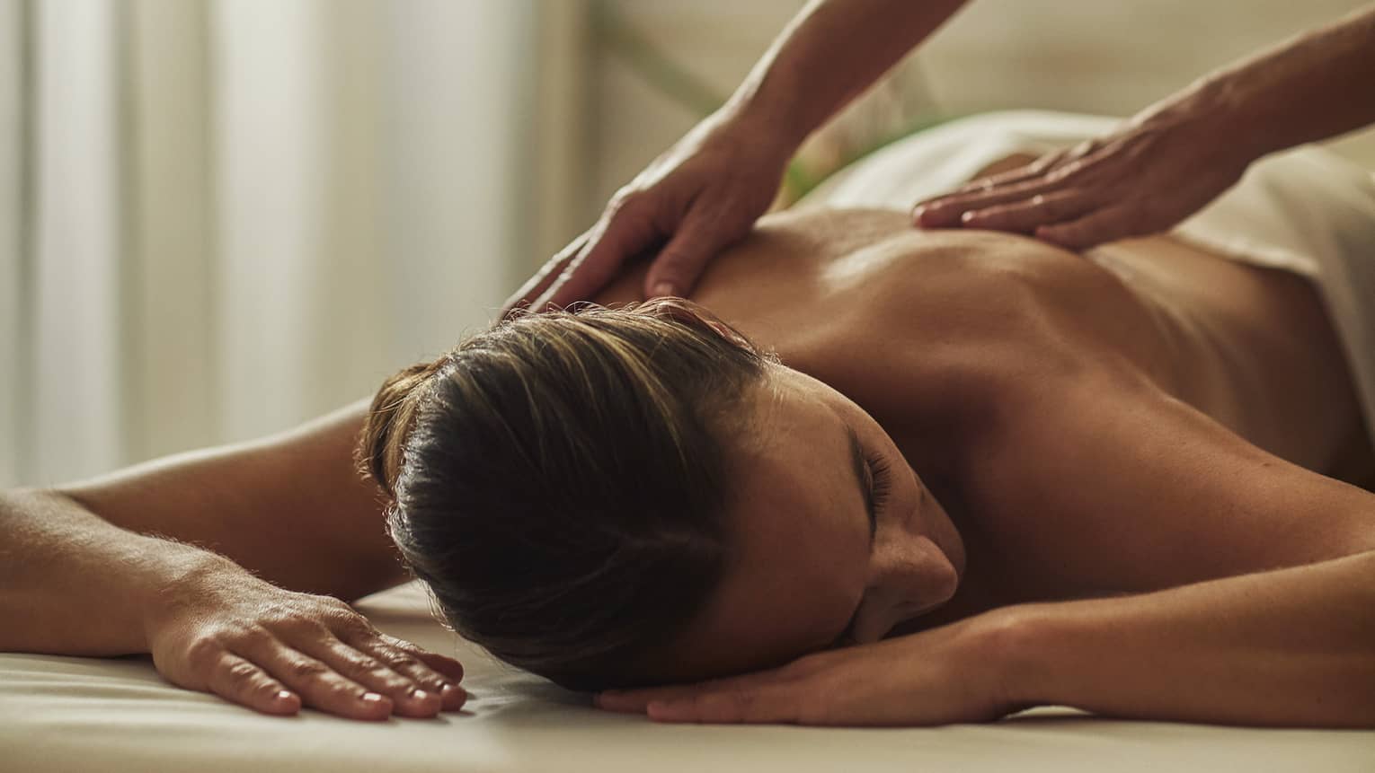 Woman in black uniform massages bare shoulders of woman lying on table face-down with head to side