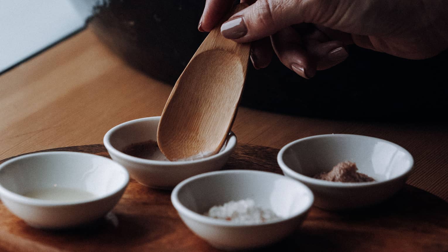 A hand places a wooden spoon into a small clay bowl to scoop up a spa ingredient, with three other clay bowls placed nearby