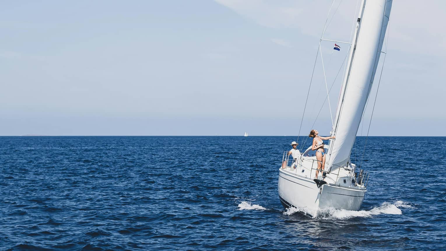 Front view of two guests aboard a sailboat as it leans into an expanse of dark blue waters under an azure cloudless sky.