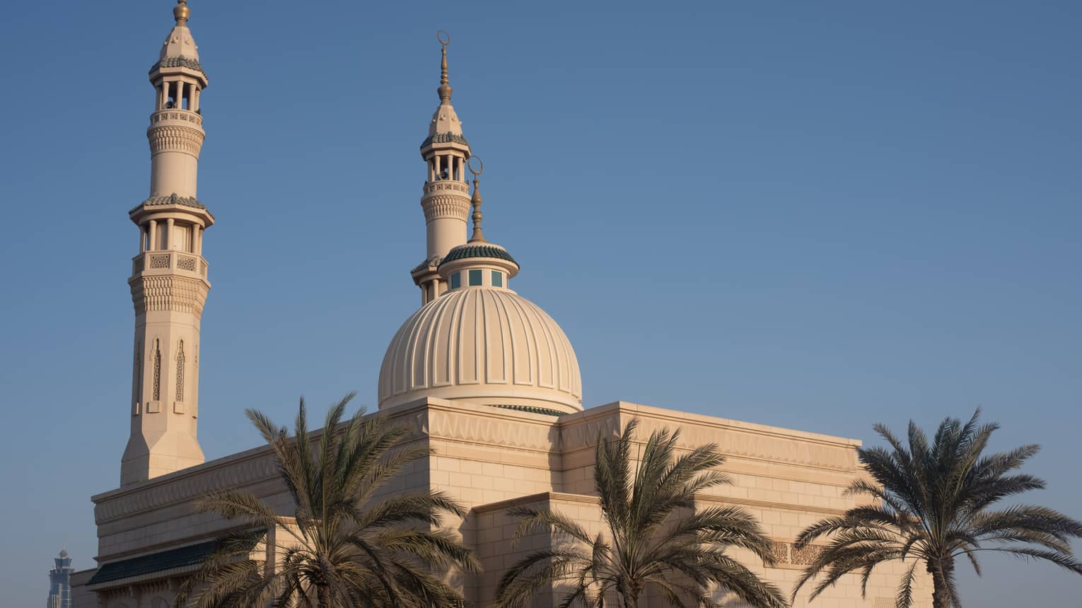A trio of palm treetops against pale sandstone of a modern mosque, two minarets and a dome reaching toward the azure sky.