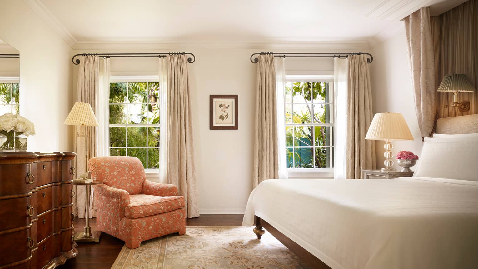 Wood dresser, pink floral armchair by tall sunny windows, nightstand and bed