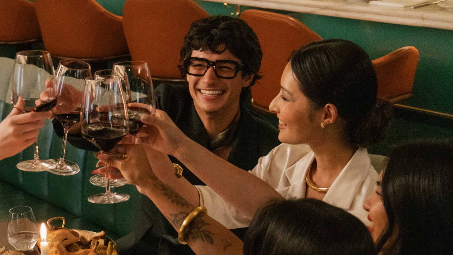 Diners seated in a banquette smile as they clink their red-wine glasses in a toast to a sumptuous candlelit feast.