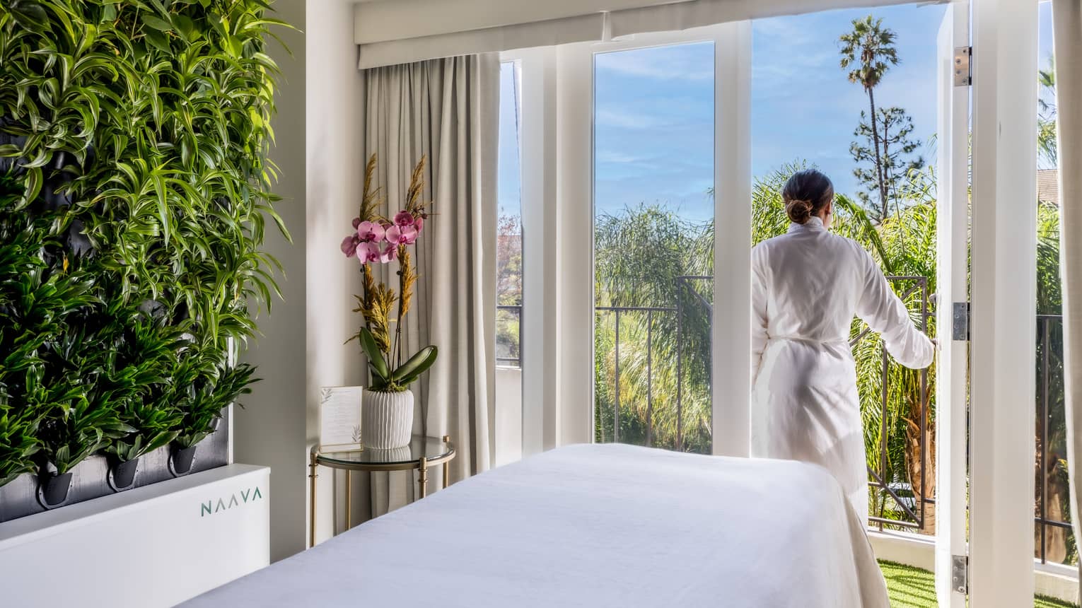 A woman walking outside from a massage room with large windowed doors and plants on one wall.