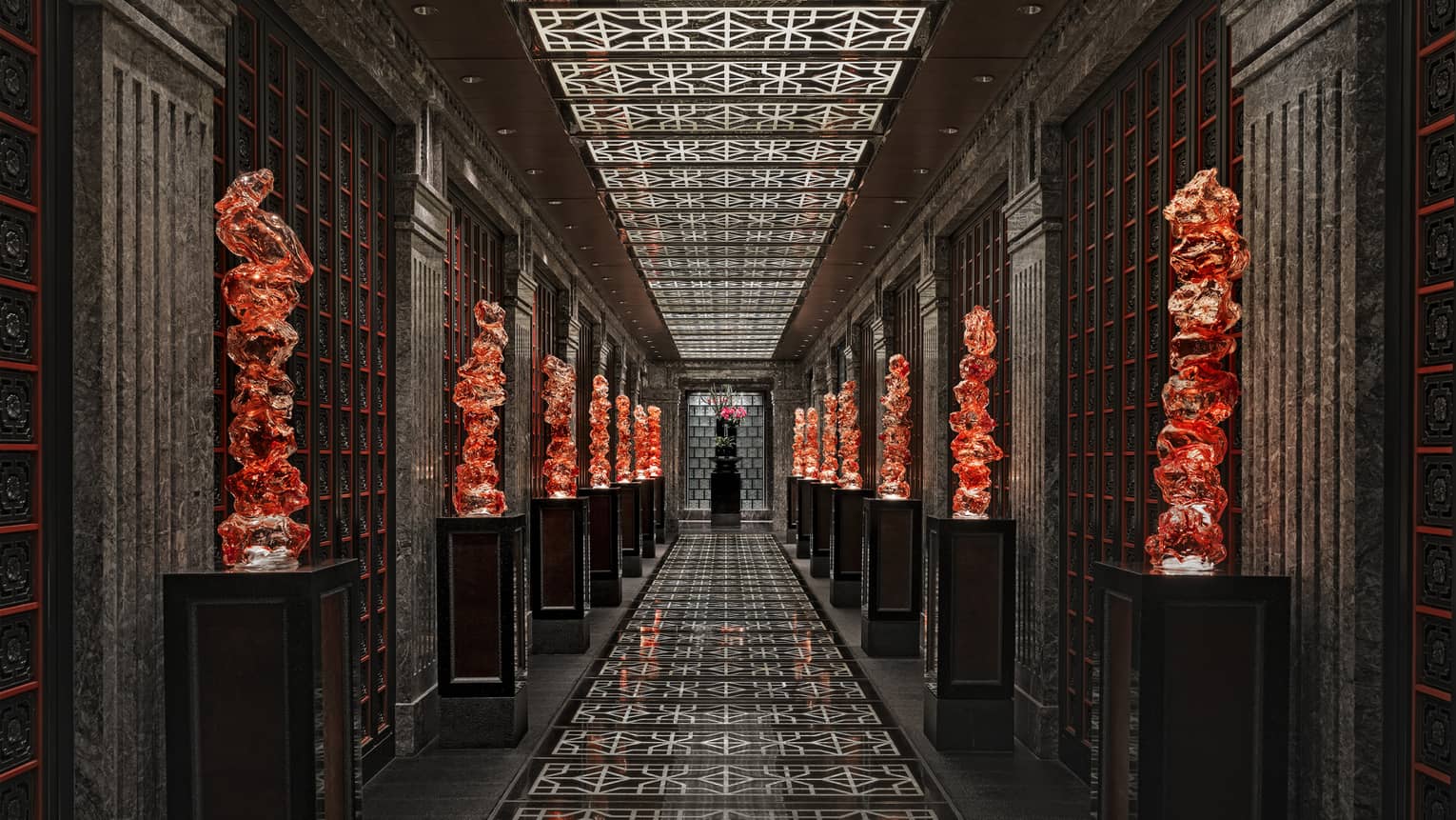 View down a long black-tiled hall lined with orange glass sculptures on pedestals, the geometric ceiling mirroring the floor.