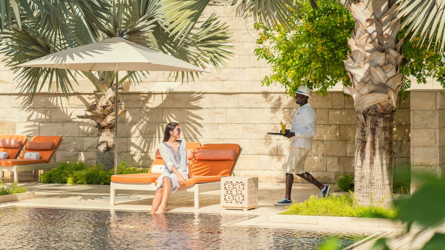 A woman lounges on an orange chaise in front of a rectangular pool as a waiter brings her a drink on a platter.