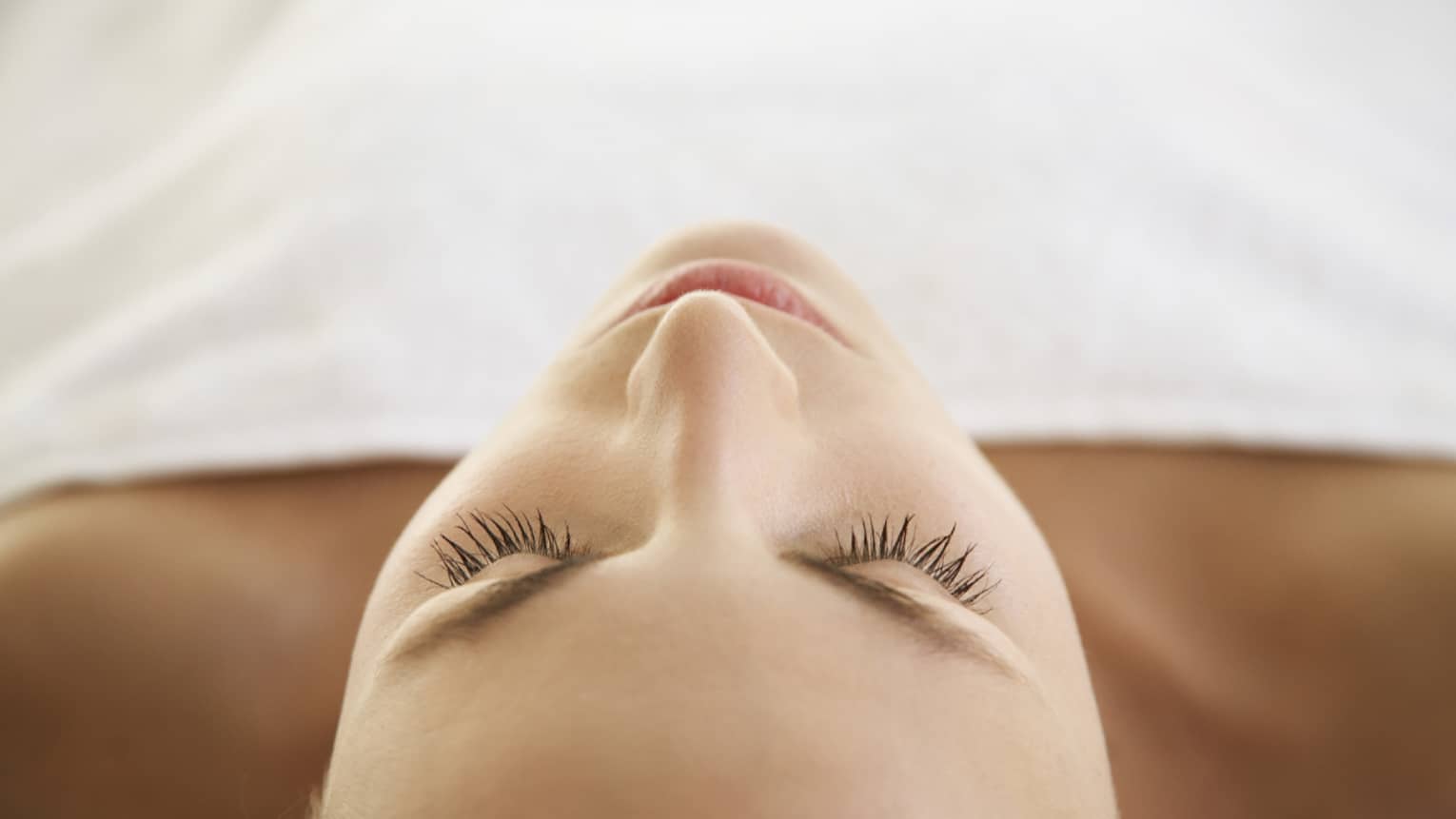 Woman lying down with eyes closed, bare shoulders, covered with white sheet
