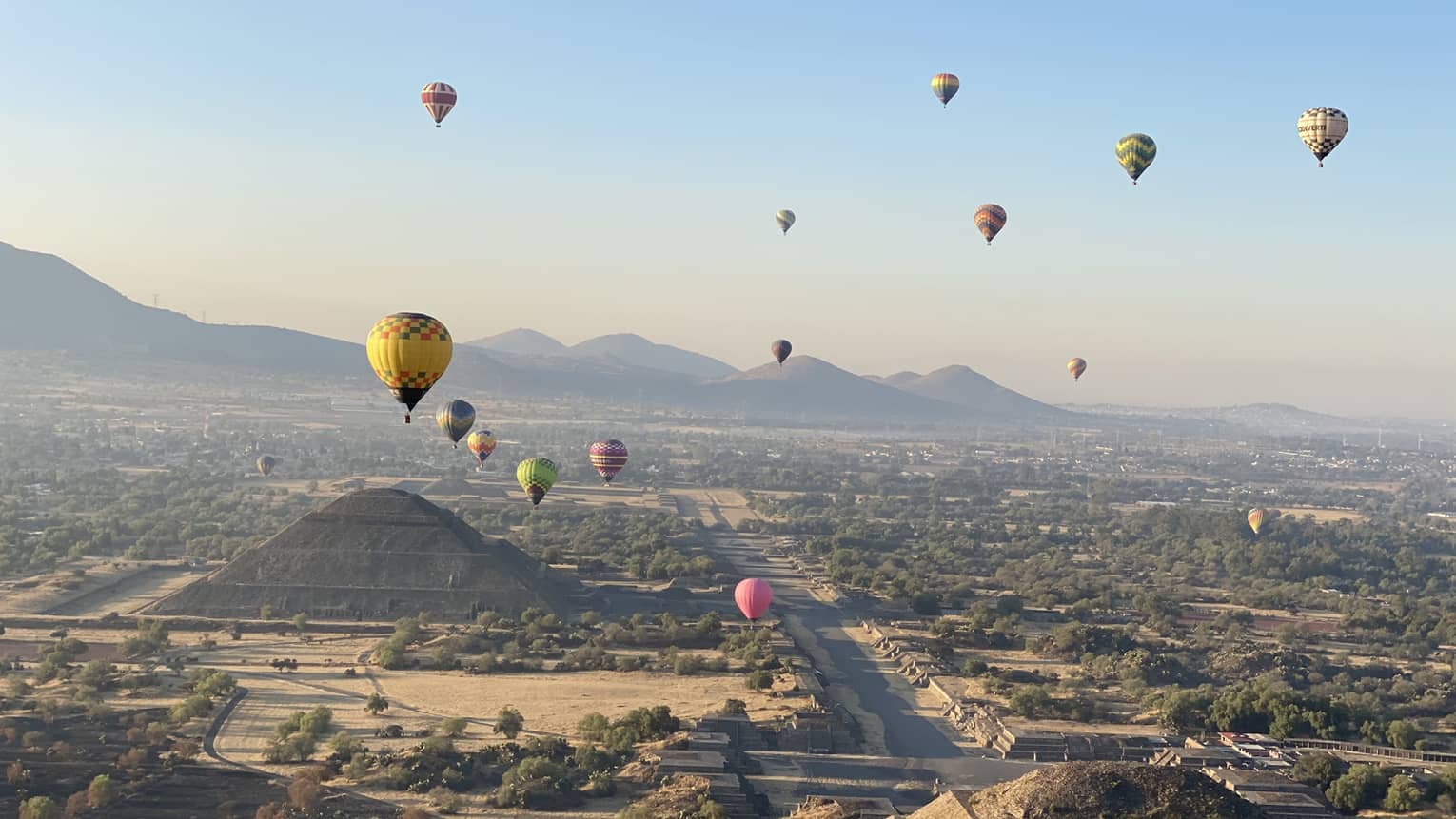 An aerial view of colourful hot-air balloons floating over pyramids and trees, with mountains and a city in the background.