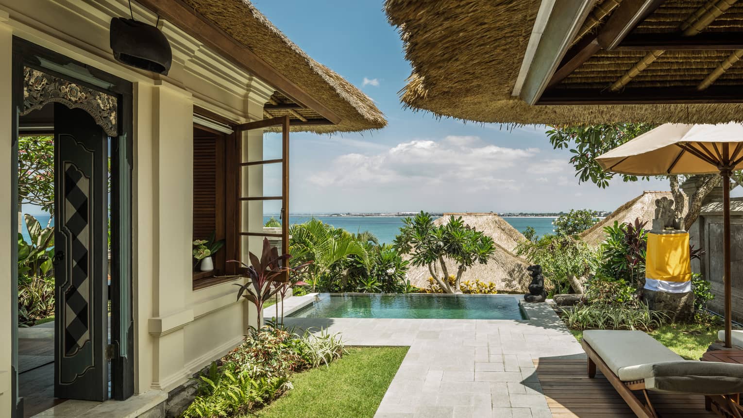 Pathway between white villa exterior wall and thatched-roof patio, with pool, palms, roofs and ocean in view