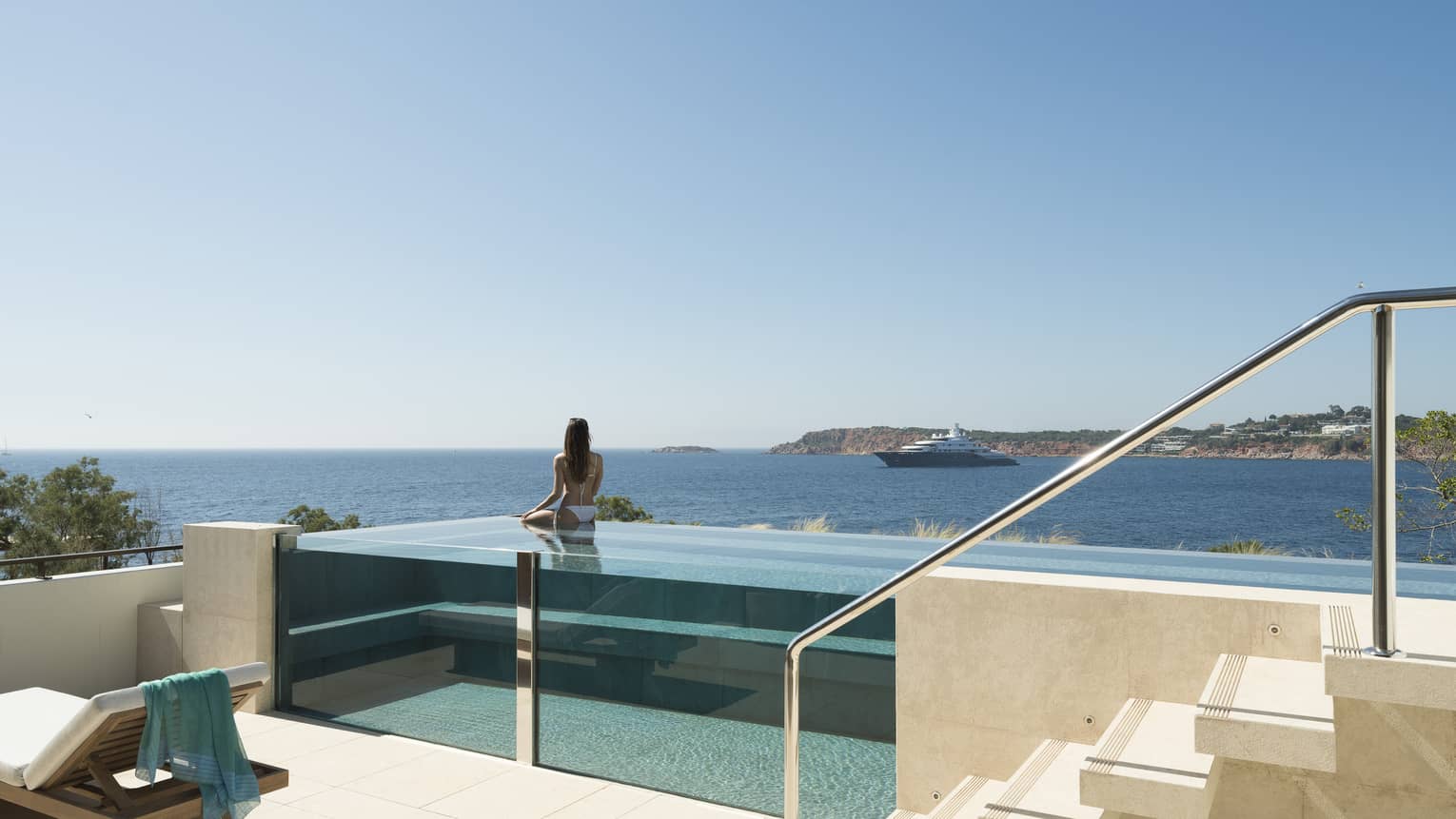 Terrace with staircase and glass infinity pool, a woman wearing white bikini in pool looks out to sea