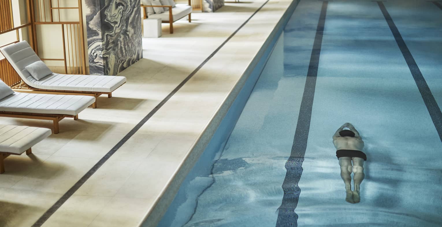 Man swims underwater in lane of indoor lap swimming pool by pool deck, chairs, marble wall