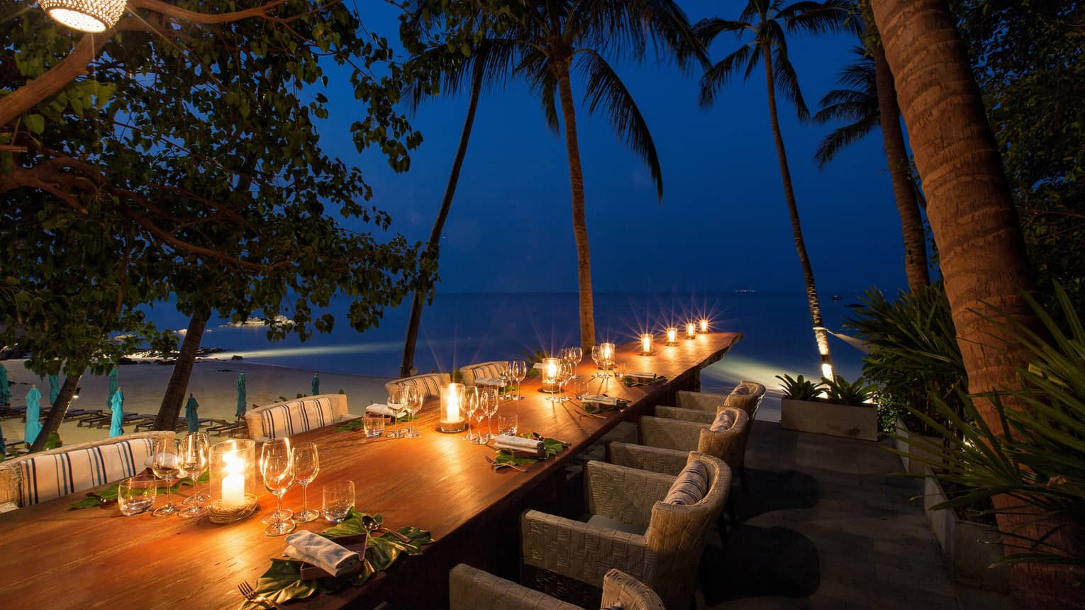 Long wood outdoor dining table with glowing candles under palm trees at night