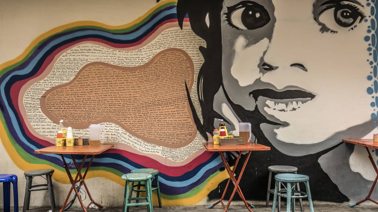 Street art mural featuring a large black-and-white face on a wall, with small, colourful dining tables and stools in front it