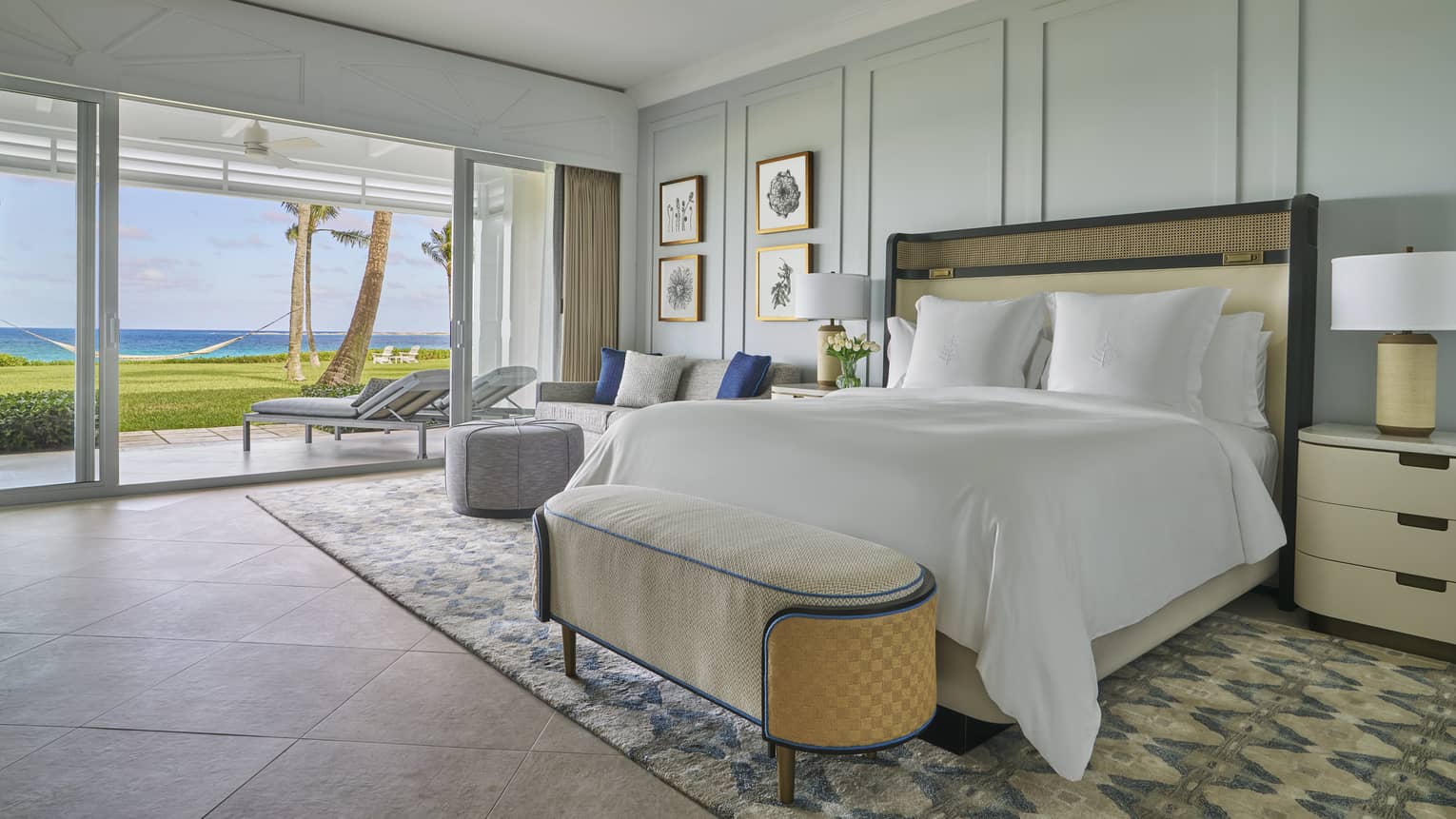 Luxury oceanfront bedroom with a large bed, cozy seating and sliding glass doors opening to a private patio with lounge chairs 