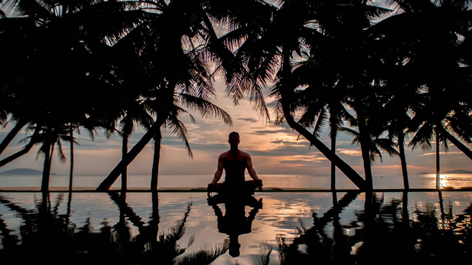 A man sitting in a yoga position at sunset next to an infinity pool surrounded by shadowy palm trees and vast ocean views