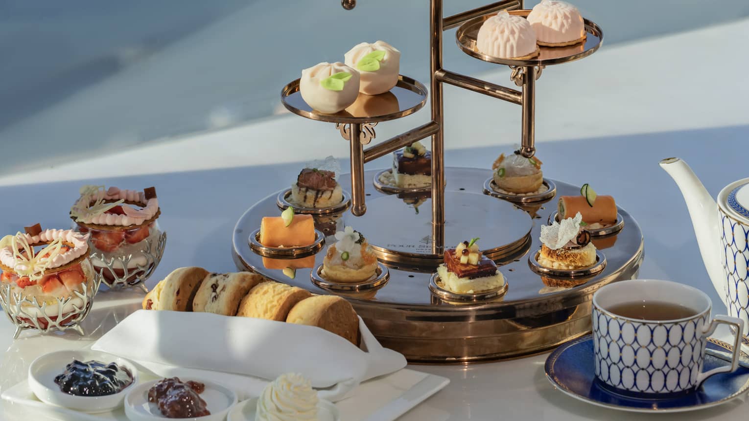 Close-up of a teacup, scones, tiny plates of jam and clotted cream, and a glossy tiered tray laden with delicate sweets.