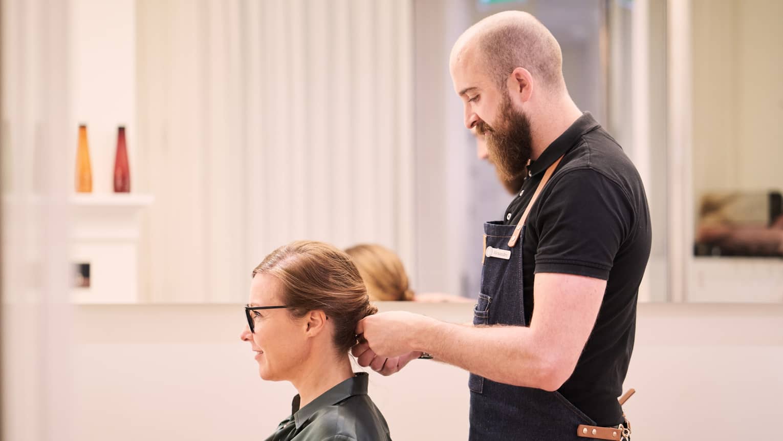 Woman sits in salon chair as hair stylist with long beard holds her hair