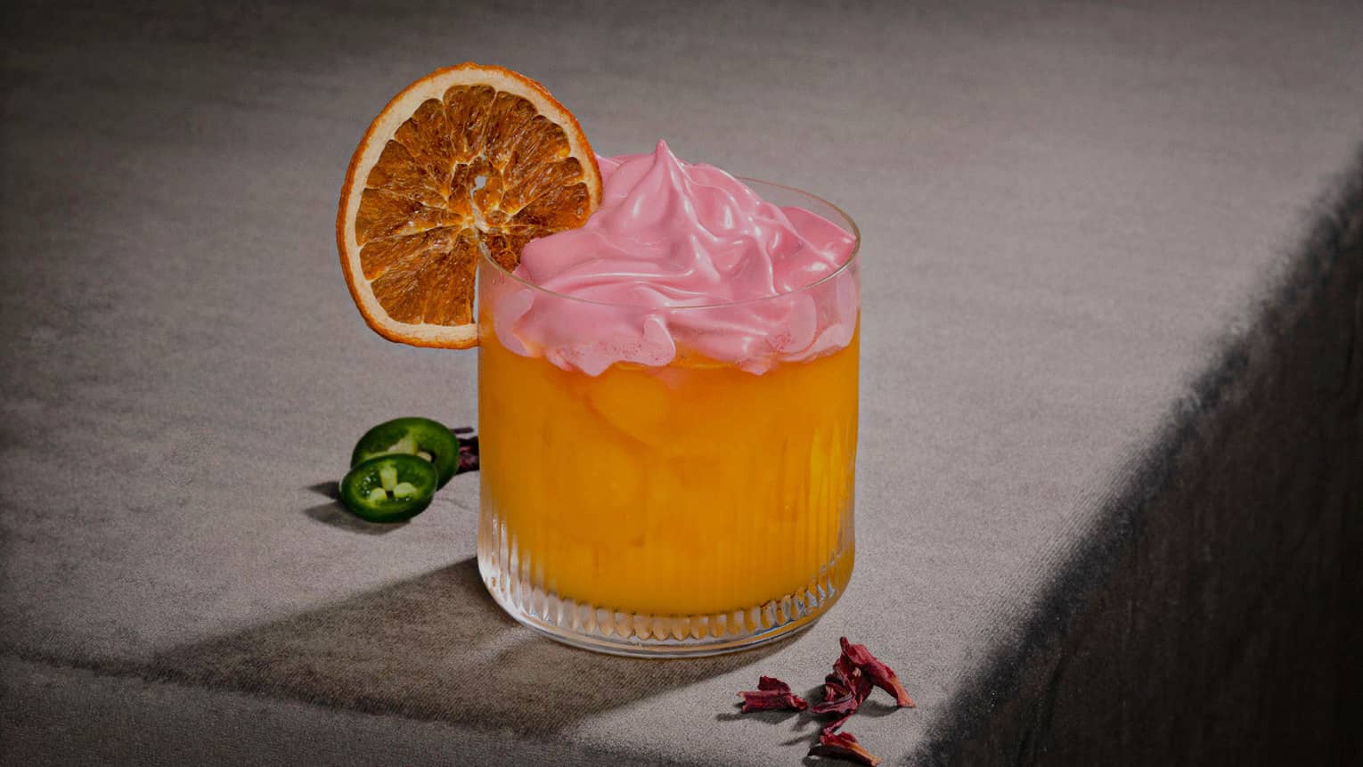 Orange-colored Mal de Ojo cocktail in a lowball glass topped with pink-hued cream and a dried orange slice on the rim