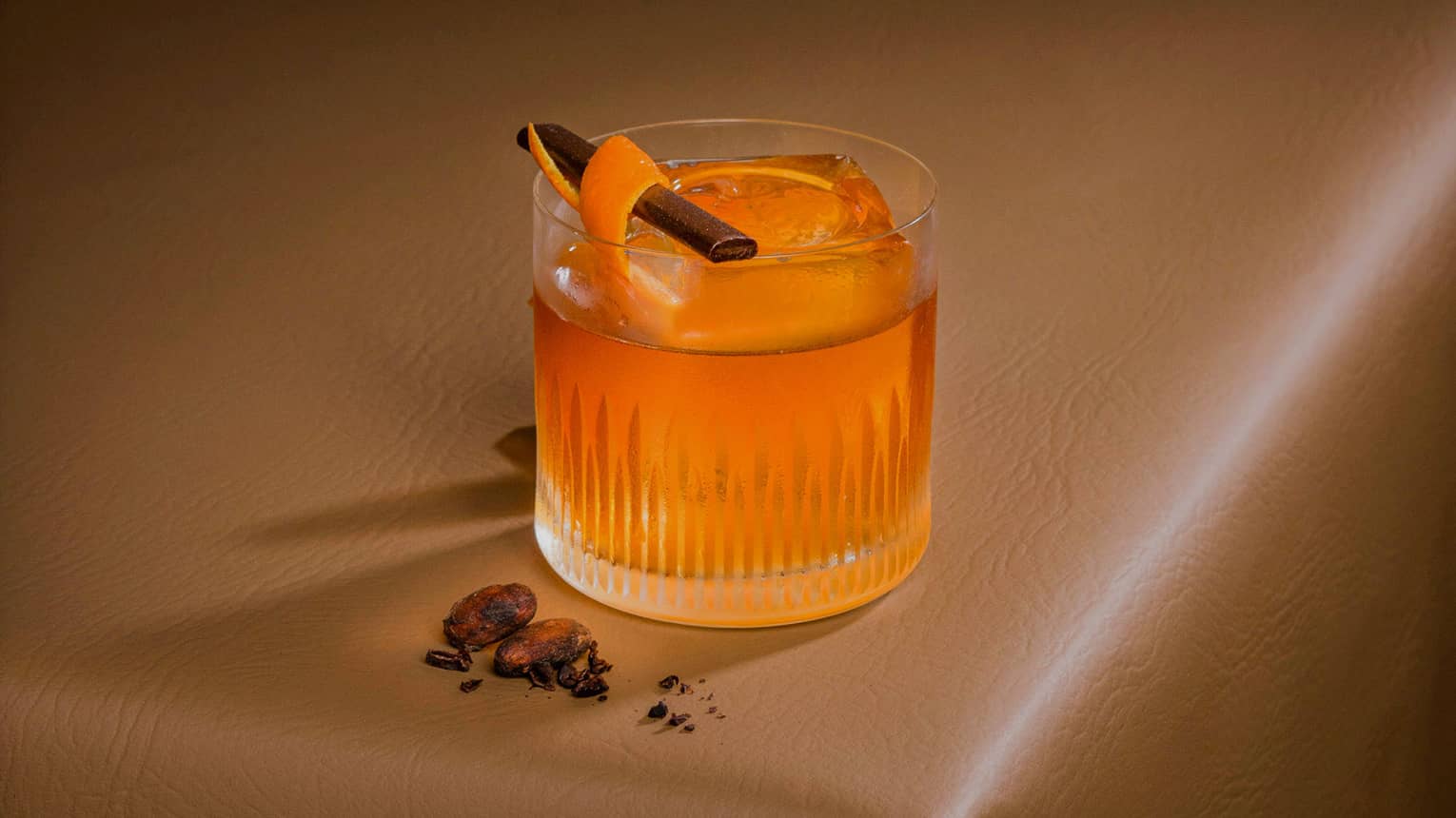 Segua's Old Fashioned cocktail in a lowball glass garnished with an orange peel and cinnamon stick sits on a brown leather tabletop